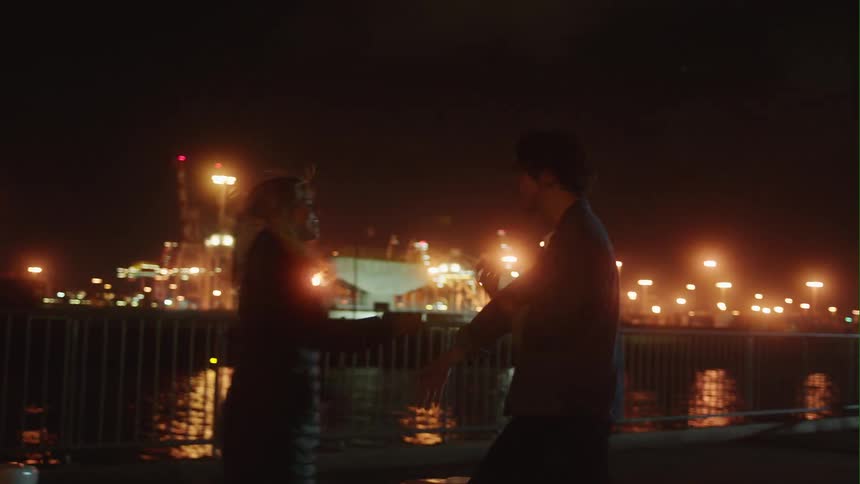 A man and a woman are standing on a boardwalk at night fighting and she pushes him and then they hug