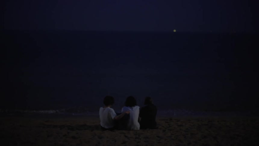 One person places jacket around anothers shoulders as three people sit together on sand looking toward ocean