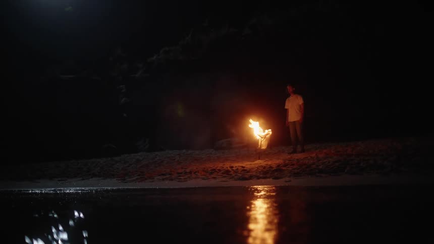 A man standing on the sand watching a fire burn and the reflection on the water surface