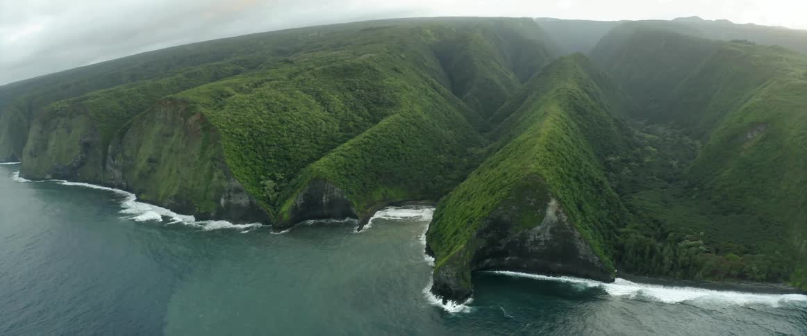 Waves roll in below grass covered mountains with coves along the coast