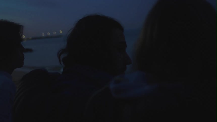 Man with long hair sits between blonde woman and man on the beach watching sea at dusk