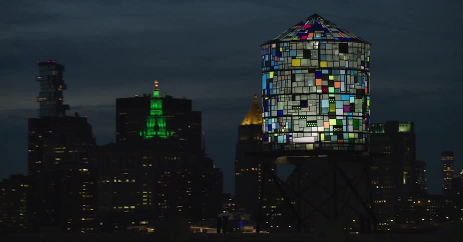 A water tower with a unique mosaic pattern stands near a metropolitan city skyline