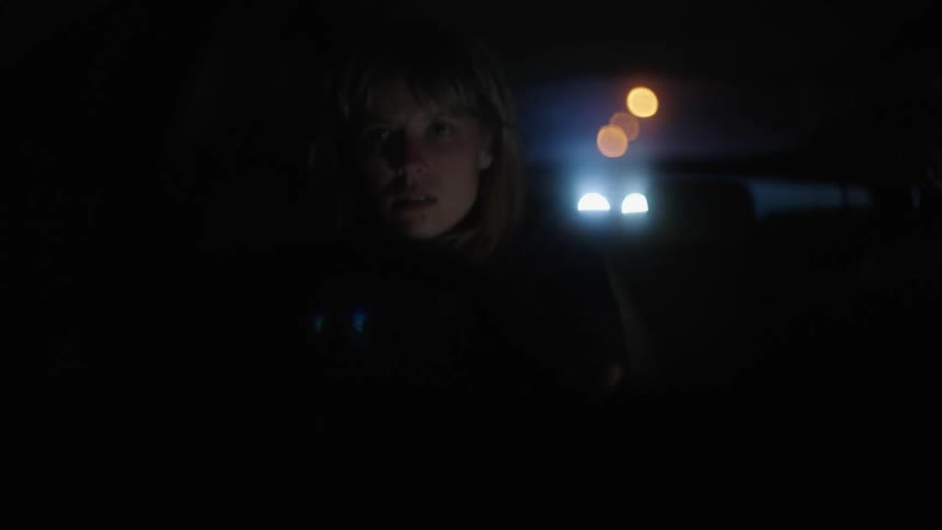 A woman sitting in the passenger seat of a traveling automobile at night wipes her face with her hand and the headlights of the vehicle in the rear are shining