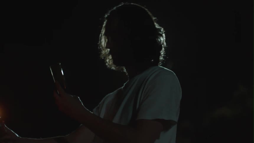 Man holding smartphone and lit lighter bends down to light fabric in field