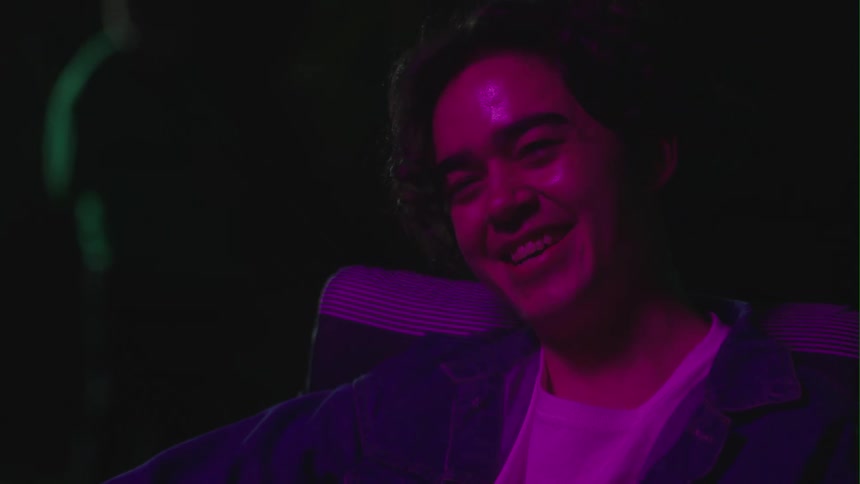 Man with denim jacket and white shirt sits in armchair drinking beer answers phone call in purple light