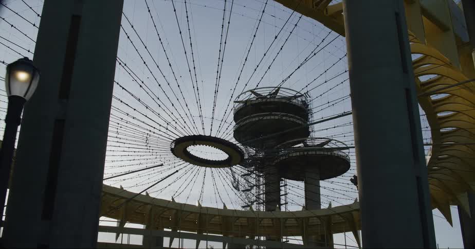 Structure designed with a circular ring and cables stands near two towers under a blue sky