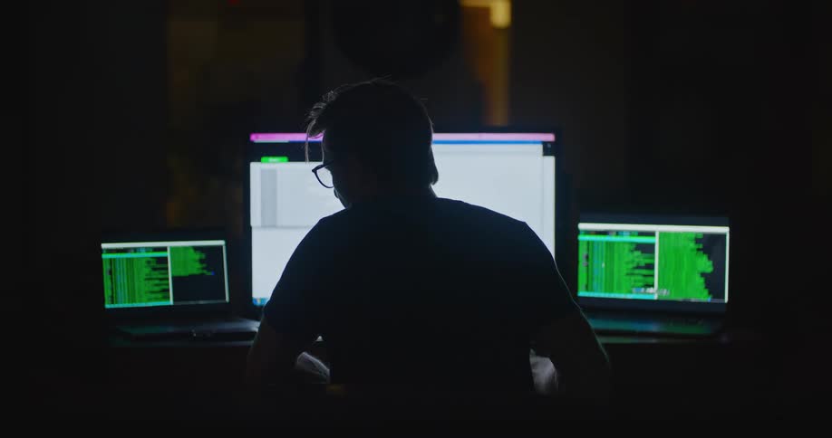 A man sits in front of three computer screens while reviewing many lines of information in the dark