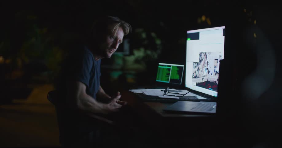 A man in a dark office throws down his pen and takes his glasses off and looks away from his computer before looking back at it