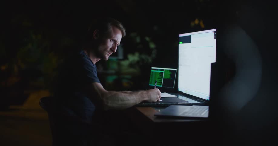 Man writes while working in dark room at desk with laptop and two other display devices
