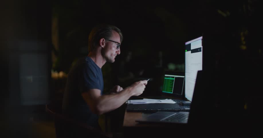 A man sitting in a dark office rubs his face as he thinks while looking at two computers