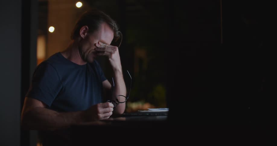 A man is sitting in a dark room at a desk with computers and talking on the phone as he looks back and forth at the computers
