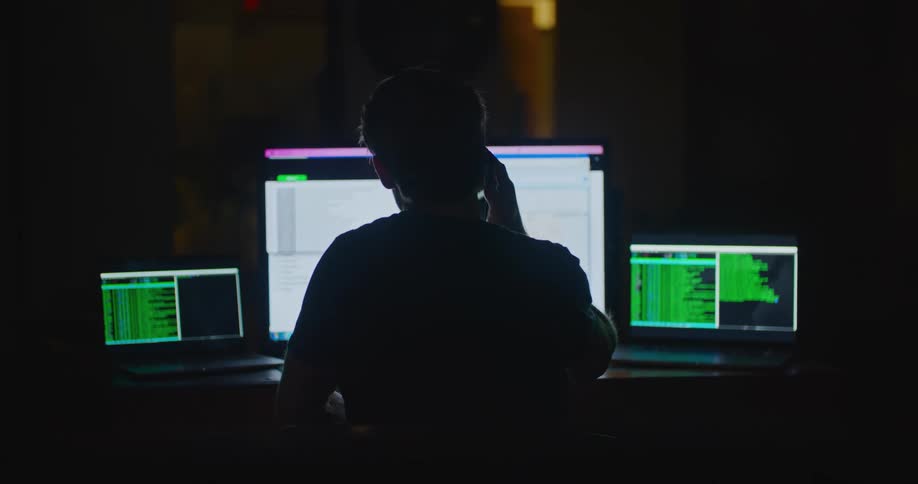 A man sitting in a dark office looks at his computer and writes on paper and then picks up his cell phone to look at it before working on a computer again