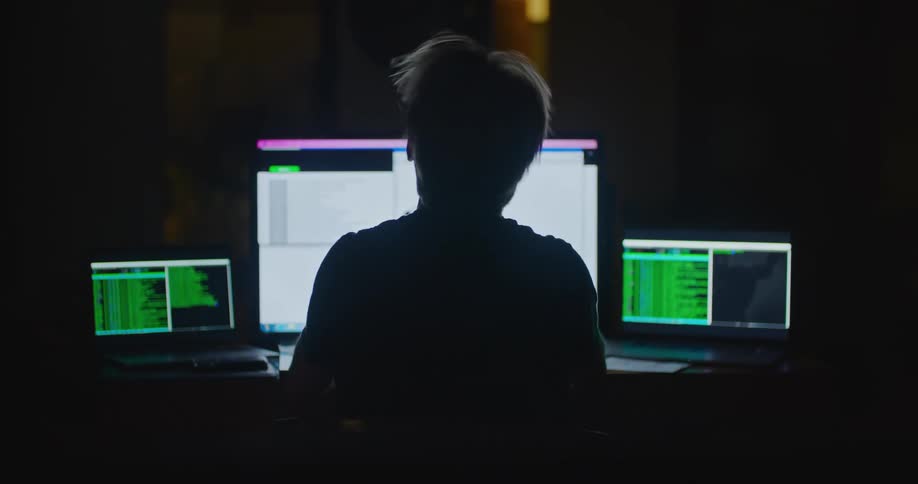 A man sitting in a dark office looks at his computer writes a note and takes off his glasses and rubs his forehead