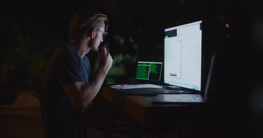 Focused middle aged blonde man wearing glasses and dark blue shirt sits at desk working on lapop and computer in dark room