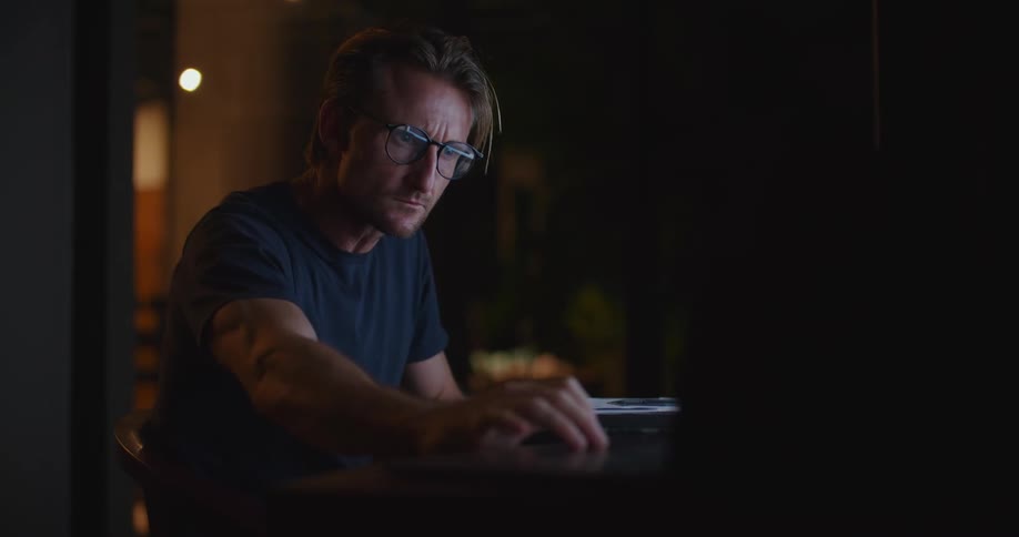 Middle aged blonde man wearing glasses and dark blue t shirt works on computer and takes notes in dark room