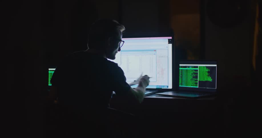 Man sits working at table in dark room with three computers