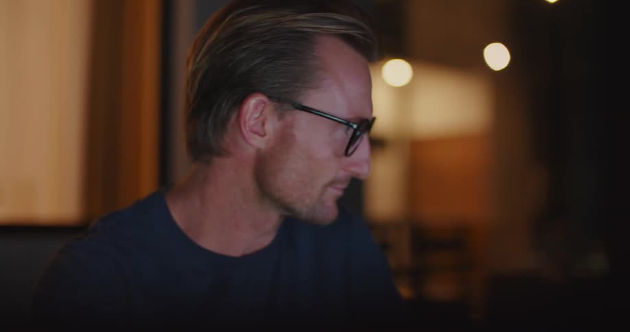 Man smiles slightly and works using computer reflected in his eyeglasses at night