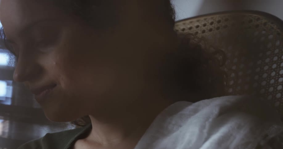 Mother with curly hair sits in swinging chair looking down and smiling next to window
