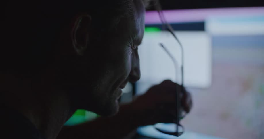 A man studying maps on a computer monitor in a dark room takes off his glasses to rub his eyes