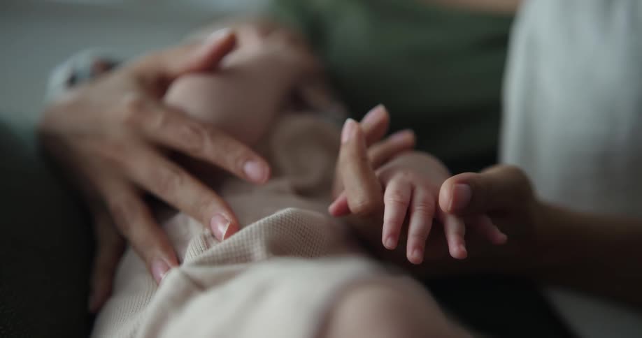 Mother caresses the hand of his newborn baby while sitting on a chair at home