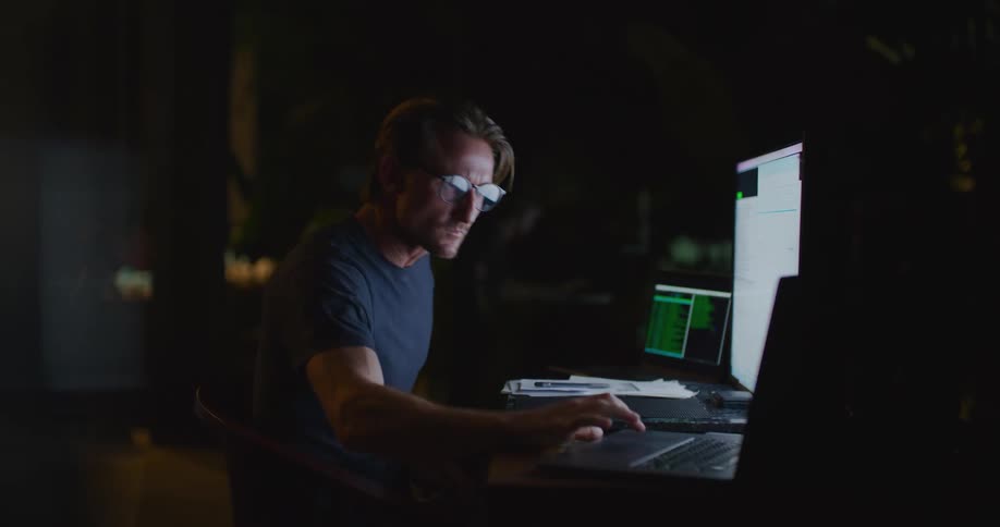 A man rubs his eyes and gets back to work looking at a monitor and typing on a laptop