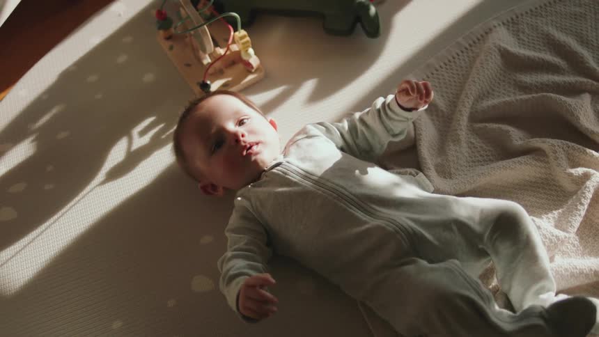 A baby is lying down kicking and moving his arms as sunlight shines on his face