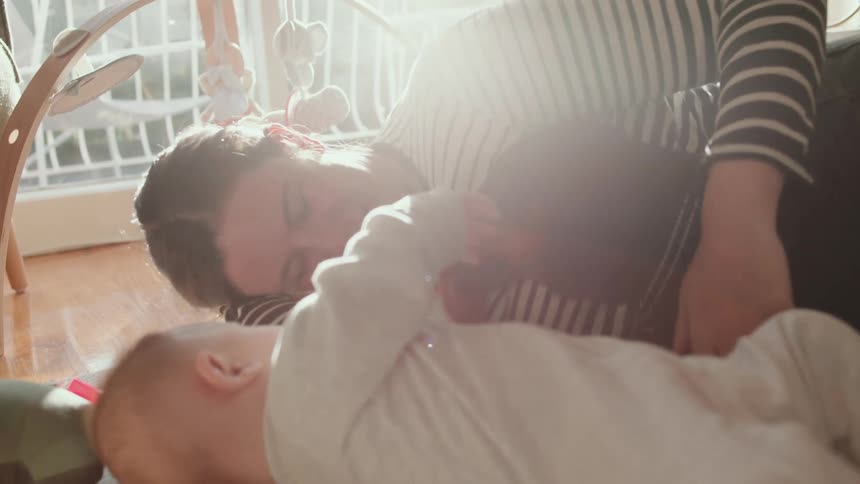Woman with striped shirt and grey jeans lies on ground cuddling brown dog next to baby in living room at home