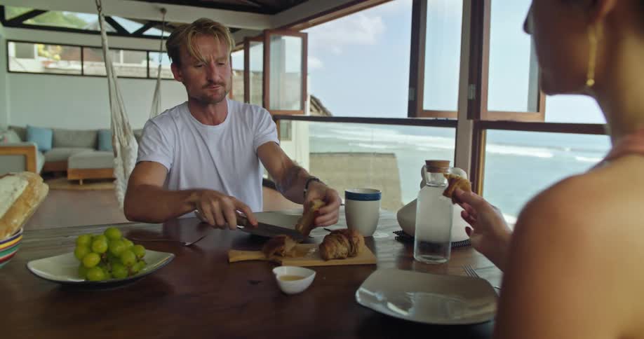 Middle aged blonde man with tattoo and white t shirt cuts meat sitting at table with woman wearing earring and pink top eating