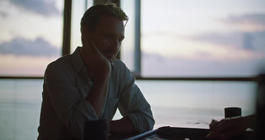 A man is sitting near a window overlooking the water playing a board game with another person