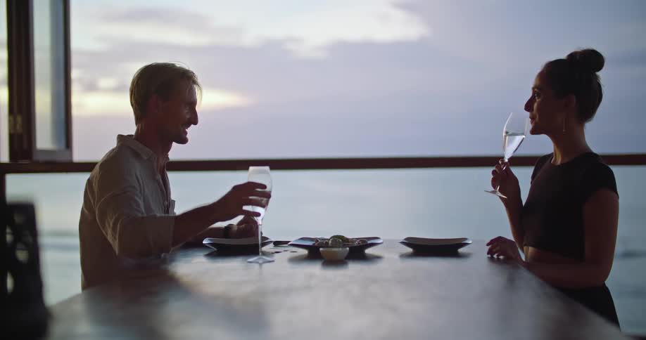 A man and woman sitting together at a dining table raise their glasses in a toast before drinking