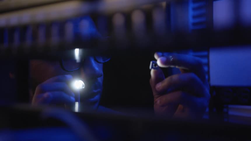 A man wearing glasses raises a little component and shines a small flashlight on it