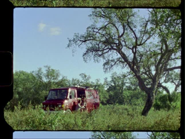 Young couple sitting on the door of a van parked in a field near a tree caught on vintage film