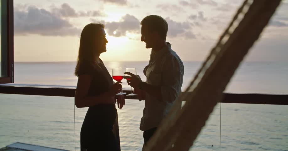 Couple smile and clink wine glasses as they talk at rail near the ocean with the sun partially silhouetting them from the horizon