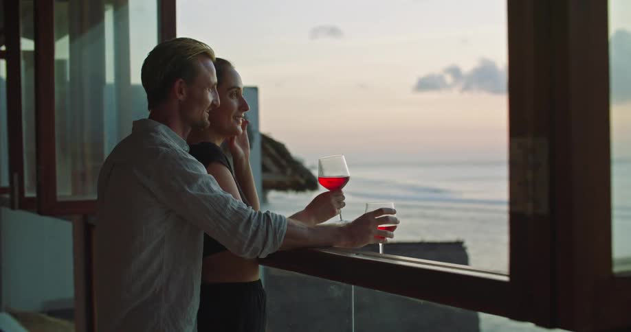 A man and woman stand at a window with wine as they look upon the ocean