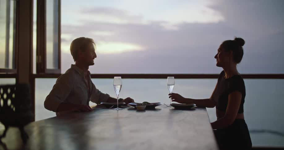 Middle aged blonde man brings a plate of food to table and clinks wine glasses with woman with bun and dress in room looking over the sea at dusk