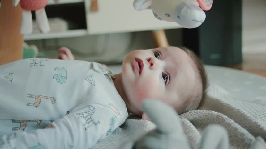 A baby lies under hanging toys on a blanket at home