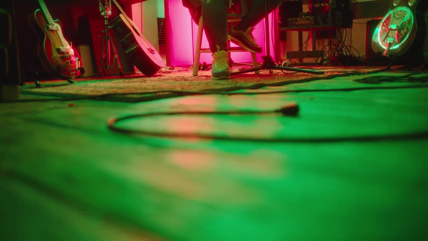 Colored lighting tints surfaces and guitars green and pink as person sits on stool while tapping their foot