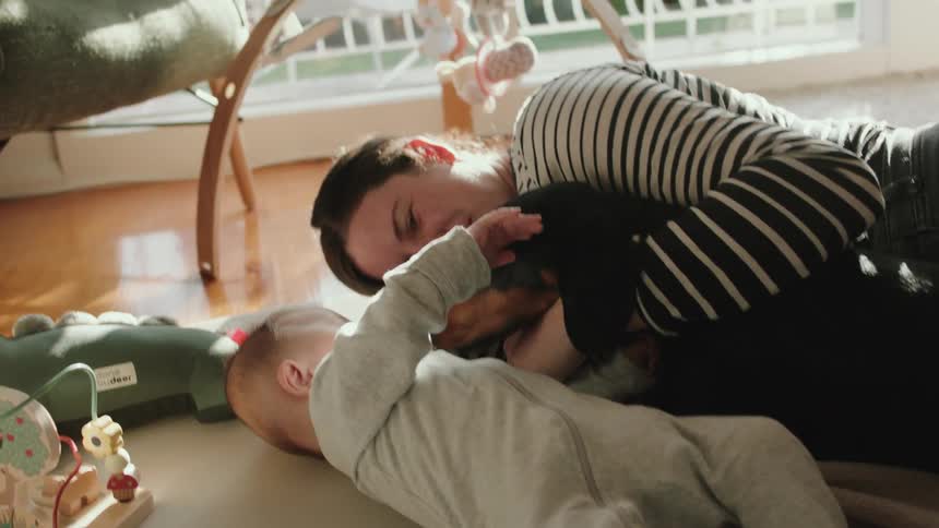 Woman with striped shirt and grey jeans lies on ground cuddling brown dog next to baby in living room at home