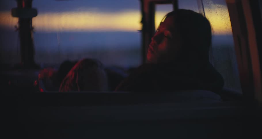 An asian woman relaxes in a parked car, rain runs down the windscreen as the sun sets