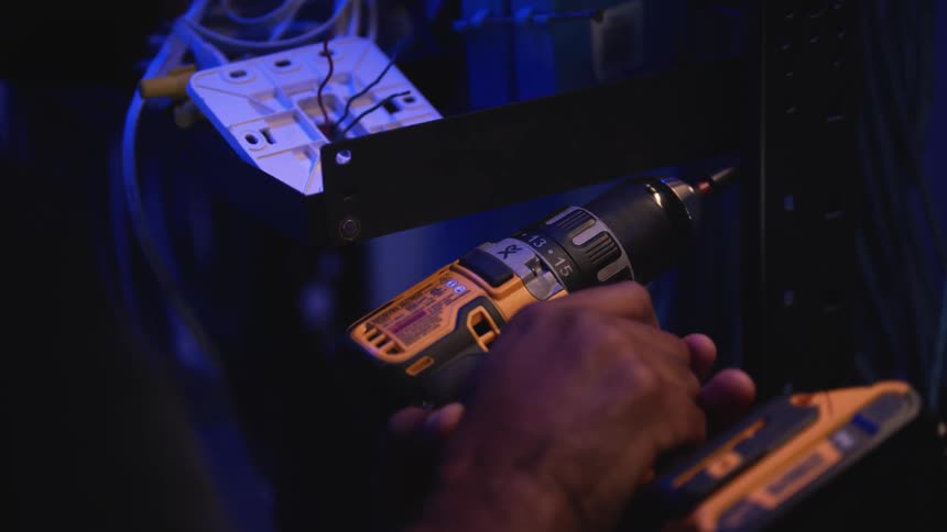 A man is working in a dimly lit room using a hand drill to tighten screws in a rack