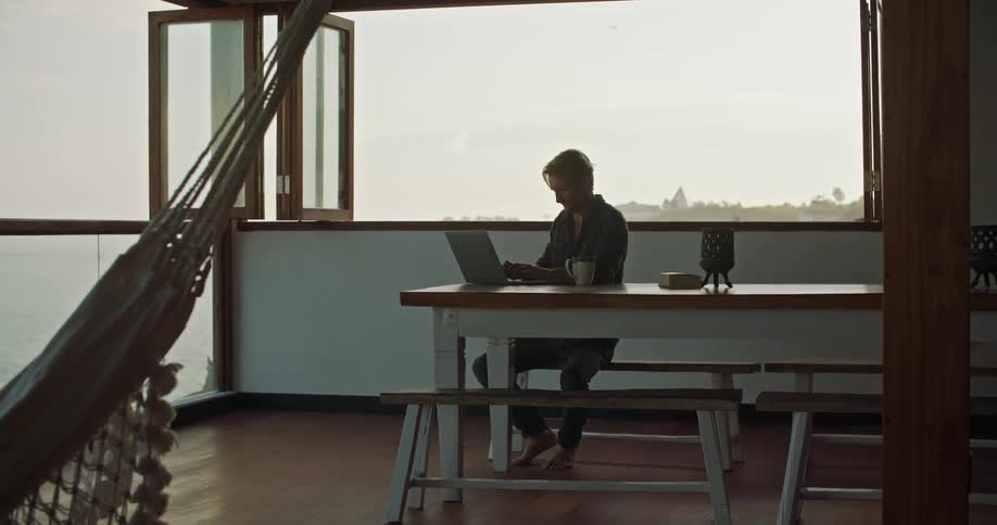 A man types on a laptop while sitting indoors at a table near a large open window during the day