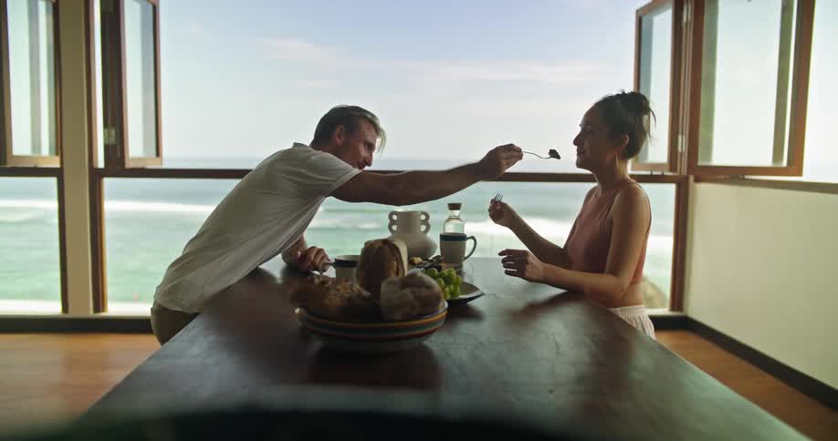 Middle aged blonde man with tattoo and white t shirt sitting at table with woman wearing earring and pink top eating food in room looking over the sea