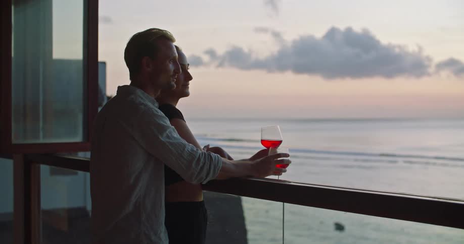Young couple drinking red wine reclined on a window looking at the ocean at sunset