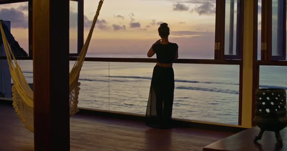 A woman wearing an evening dress is taking a picture of the waves rolling into shore outside the window