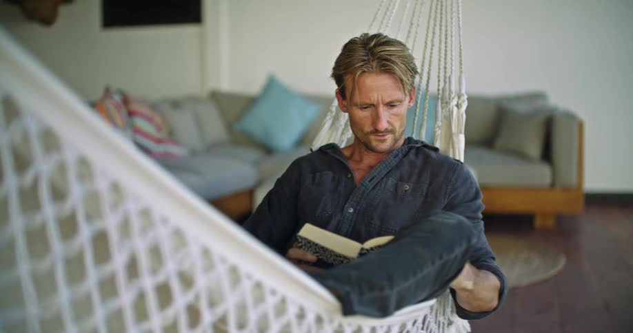 Middle aged blonde man with dark blue shirt and jeans sits in hammock reading a book in room