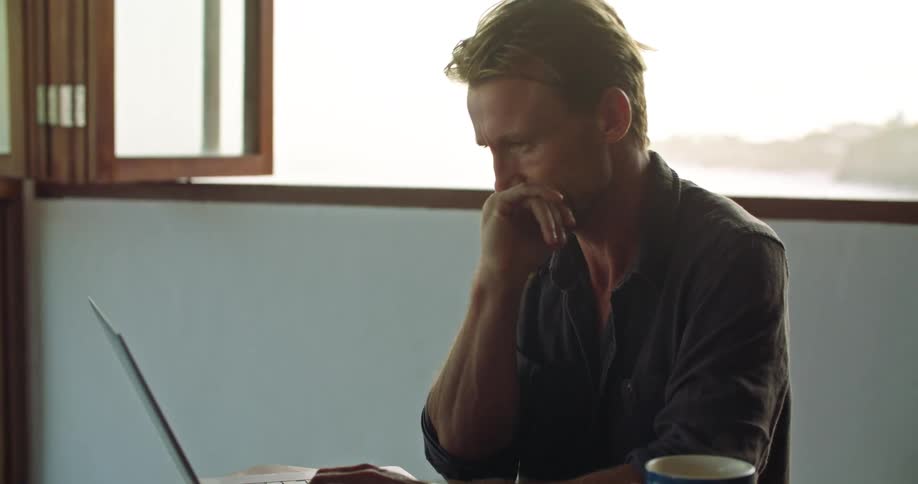 Middle aged blonde man with dark shirt sits at table working on laptop in room
