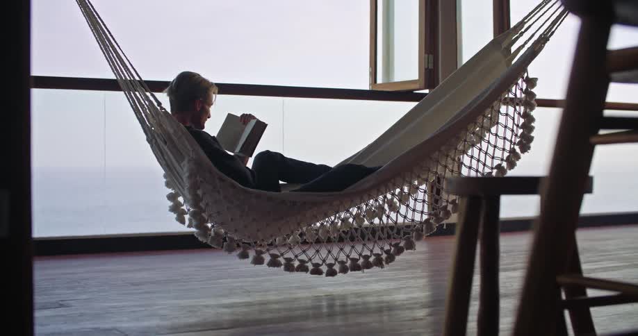 Man leisurely relaxing over a hammock while reading a book inside an apartment