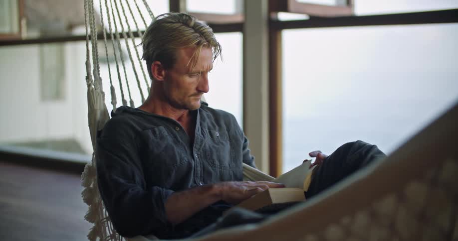 A man is reading a book and sitting in a hammock
