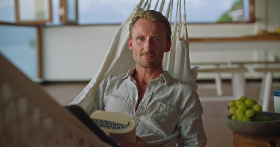A man is sitting on a hammock holding a book and he smiles as he gently rocks back and forth