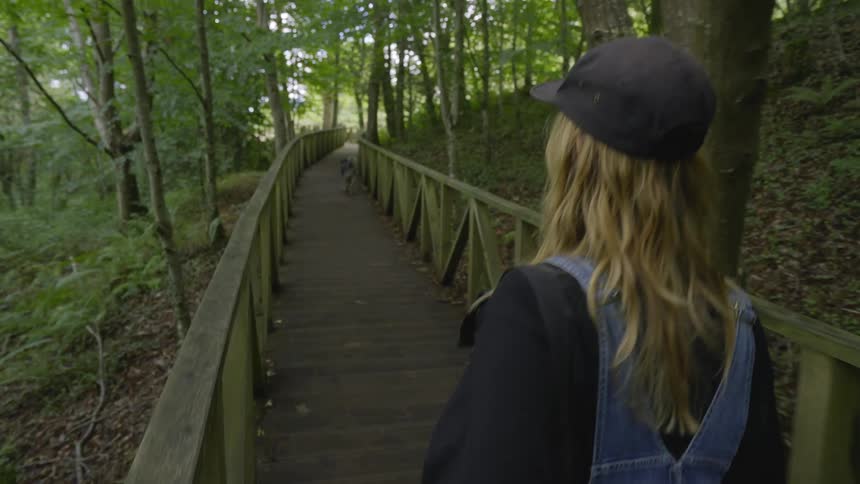 Young blonde woman walking on a wooden deck through the forest with her dog